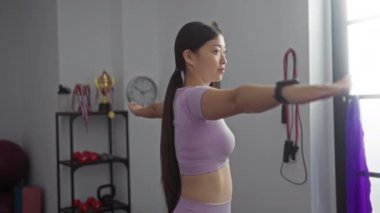 Spor malzemeleri ve ödüllerle dolu bir spor salonunda kollarını açmış egzersiz yapan genç bir kadın..