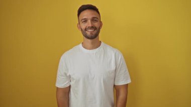 Beyaz tişört giyen genç İspanyol adam sarılmak için kollarını açmış neşeli bir ifade, mutluluk saçan, izole edilmiş sarı arka planda kameraya gülümseyen.