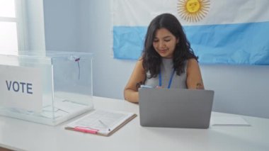 Arjantin 'de seçmen odasında cep telefonu kullanan genç bir kadın. Arka planda oy sandığı ve bayrak var.
