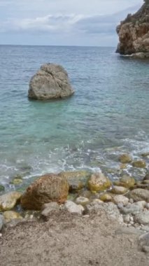 Mallorca 'da Cala Deia' nın kayalık kıyı şeridi. Turkuvaz suları çakıl taşlarını okşuyor ve bulutlu bir gökyüzünün altında durmakta olan bir kaya kütlesi.