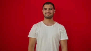 Beyaz tişört giyen genç İspanyol adam başparmağıyla dikiliyor, mutlu görünüyor ve izole edilmiş kırmızı arka planda gülümsüyor.