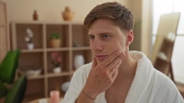 Beyaz bir bornoz giyen ve yüzüne dokunan genç adam düşünceli, yeşil bitkiler ve yumuşak ışıklandırma ile kapalı bir sağlık merkezi