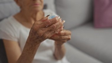 Kısa saçlı, olgun elleri olan yaşlı bir İspanyol kadın elinde bir şişe hapla oturma odasında sakin ve kapalı bir atmosferde oturuyordu..