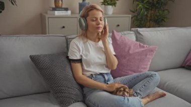 Genç sarışın kadın, sıcak bir oturma odasında Chihuahua köpeğiyle kulaklıkla müzik dinliyor..