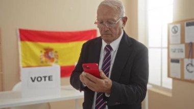 Beyaz gözlüklü yaşlı bir adam akıllı telefon kullanıyor, kapalı bir oylama odasında İspanyol bayrağı ve oy kutusuyla duruyor, İspanya 'daki seçim sürecini temsil ediyor..