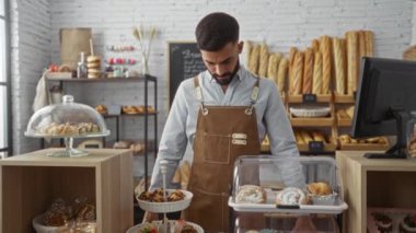 Önlüklü genç adam kollarını kavuşturup raflarda çeşitli hamur işleri ve ekmeklerle dolu bir pastanede gülümsüyor..