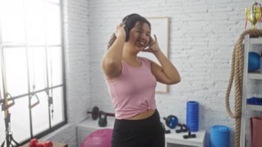 Spor salonunda kulaklıklarla dans eden genç bir kadın, spor aletleri ve spor ekipmanlarıyla çevrili, kapalı bir spor ortamında neşe ve enerji saçıyor.