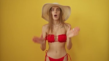Bikinili ve yaz şapkalı kadın şaşırmış ve şaşırmış bir ifadeyle izole edilmiş sarı arka planda şok olmuş ve korkmuştu.