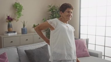 Parlak bir oturma odasında sırtı ağrıyan, kısa saçlı, iç mekan bitkileri ve minimalist dekoru olan yaşlı bir İspanyol kadın..