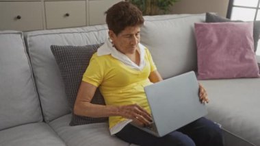 Sarı tişörtlü yaşlı bir İspanyol kadın modern oturma odasındaki rahat gri bir kanepede otururken dizüstü bilgisayar kullanıyor..