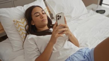 Modern yatak odasında yatakta akıllı telefon kullanan genç bir kadın ekrana bakıp esniyor ve yorgunluk belirtileri gösteriyor.