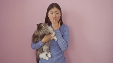 Şok olmuş genç Çinli bir kadın, şaşırmış bir ifadeyle, ağızını eliyle kapatıyor, kediyle birlikte pembe arka planda duruyor.