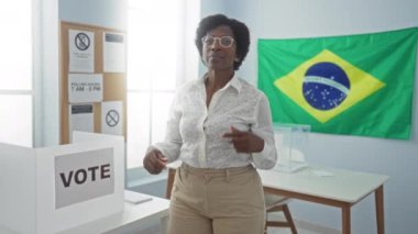 Brezilya Seçim Koleji 'nde oy sandığının yanında mutlu bir şekilde duran kadın. Orta yaşlı, Afro-Amerikan bir kadın. Gülümsüyor ve güvenle parmak ve elle işaret ediyor.