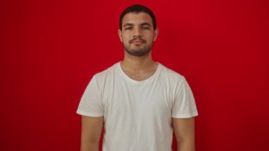 Beyaz tişörtlü genç İspanyol adam ayakta dikiliyor, işaret ediyor, şaşırarak işaret ediyor izole edilmiş kırmızı arka plan üzerinde şaşkınlık