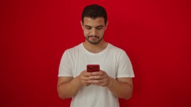 Akıllı telefonlu genç İspanyol bir adam, düşünceli bir tavır sergiliyor ve düşünceli bir ifade sergiliyor, izole edilmiş kırmızı arka planda kendine güvenini gösteriyor.
