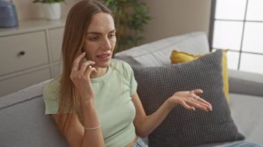 Sarı ve gri yastıklı gri bir kanepeye oturmuş yeşil mahsullü beyaz bir kadın oturma odasında telefonla konuşuyor..