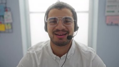 Ofis ortamında gözlüklü ve kulaklık takan İspanyol bir adam, pencerenin yanında otururken bir video görüşmesi yapıyor, profesyonel bir iş ortamını yansıtıyor..