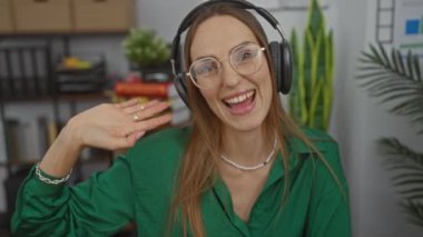 Ofiste gözlüklü ve kulaklıklı çekici beyaz bir kadın. Görüntülü görüşme sırasında seyirciyi selamlamak için elini kaldırıyor..