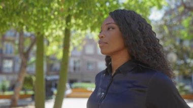 Kıvırcık saçlı Afro-Amerikalı genç bir kadın şehir parkında yürüyor, şehir hayatının sakin ve güzelliğinin bir örneğini veriyor..