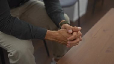 İspanyol bir adam bekleme odasında oturuyor, ellerini kucağına yapıştırıyor, düşünceli ve sabırlı görünüyor..