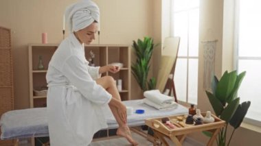 Kaplıcadaki güzellik merkezinde genç, sarışın bir kadın. Bornoz ve türban giyiyor. Masaj masasında oturuyor ve bacağına krem sürüyor..