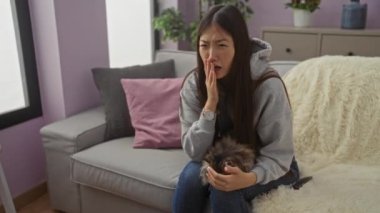 Oturma odasındaki gri bir kanepede oturan Asyalı genç bir kadın, kucağında küçük bir kedi yavrusu, sıcak bir öğleden sonra evde otururken rahatsız görünüyor..