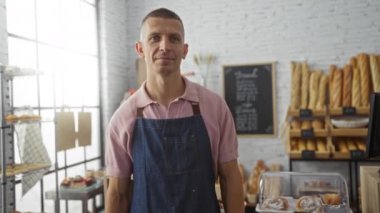 Fırın odasında genç bir adam, arka planda ekmek ve hamur işleri sergilenen pembe bir gömlek ve önlük giyerek başparmak işareti yapıyor..