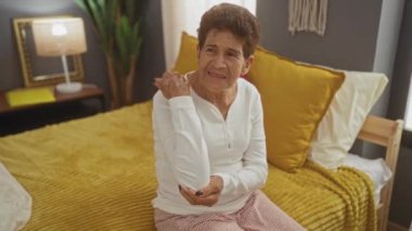 Kısa saçlı yaşlı bir İspanyol kadın rahat bir yatak odasında yatağında oturuyor, dirseğini acıdan kavrıyor, sıcak bir ışık ve ev dekorasyonu davetkar bir atmosfer yaratıyor..
