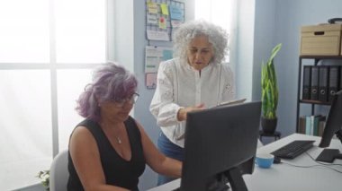 Olgun kadınlar, modern ofis içinde çalışmaktan bahsediyorlar. Bir kadın bilgisayarın başında oturuyor, diğeri ayakta duruyor ve sürekli el kol hareketi yapıyor.