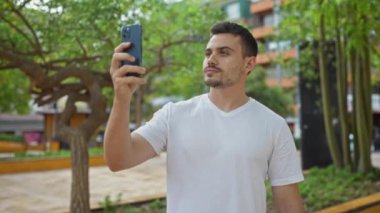 Şehir parkında sakallı yakışıklı genç bir adam dışarıda akıllı telefonuyla selfie çekiyor..