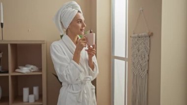 Spa merkezinde çay içen bir kadın bornoz ve havlu giymiş, arka planda mumlar ve raflarla sakin bir sağlık odasının penceresinden bakıyor.