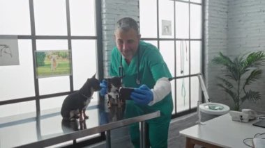 Veteriner kliniğinde orta yaşlı bir adam, etrafı tıbbi ekipmanlar ve evcil hayvan posterleriyle çevrili metal bir muayene masasında iki chihuahua ile selfie çekiyor..