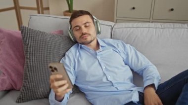 İspanyol bir adam oturma odasındaki bir kanepede dinleniyor, kulaklıkla müzik dinliyor, akıllı bir telefon tutuyor, sıradan giyinmiş ve mutlu görünüyor..