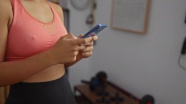 Spor salonunda genç bir kadın pembe spor sutyen giyiyor. Parlak ışıklandırılmış ev ortamında akıllı telefon kullanıyor. Spor ve teknoloji kullanımına odaklanıyor.