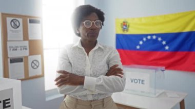 Afrika kökenli orta yaşlı, kıvırcık saçlı ve gözlüklü bir kadın seçim odasında kollarını kavuşturmuş, arkasında Venezuela bayrağı olan bir kadın..