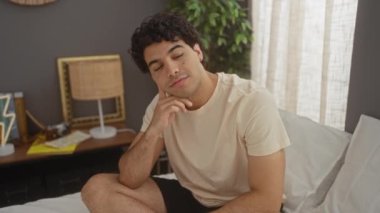 Düşünen, pijamalı, ciddi bir yüzü ve eli çenesinde yatakta oturan, kafası karışık ve düşünceli genç bir adam.