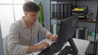 Modern bir ofiste bilgisayar başında çalışan genç bir adam düzenli raflar ve klasörlerle çevrili, profesyonel bir iç mekan ortamında verimliliği gösteriyor.