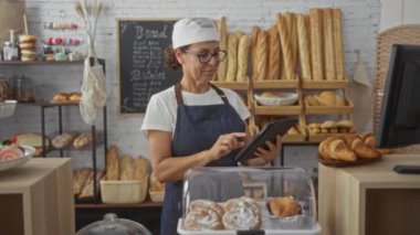 Fırında çalışan, tablet kullanan, çeşitli ekmek ve hamur işleriyle çevrili, arka planda karatahta menüsü olan bir kadın..