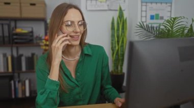 Gözlüklü ve yeşil tişörtlü çekici bir kadın modern kapalı bir ofiste telefonla konuşurken gülümsüyor ve arka planda bir raf var..