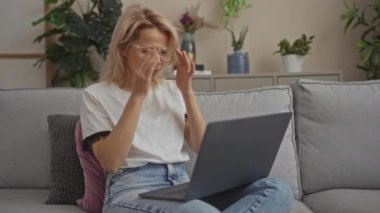 Gözlüklü genç sarışın bir kadın oturma odasındaki gri bir kanepede oturuyor. Yanında küçük köpeği olan bir dizüstü bilgisayar kullanırken stresli görünüyor..