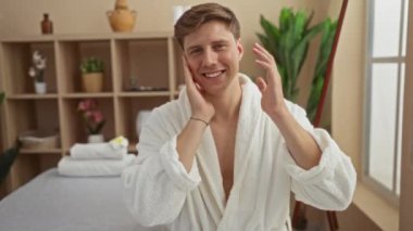 Beyaz bornozlu genç adam, çiçeklerle ve havlularla süslenmiş bir kaplıca odasında gülümsüyor ve yüzüne dokunuyor.