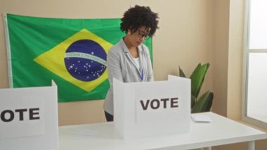 Kıvırcık saçlı Afro-Amerikalı genç bir kadın, arka planda Brezilya bayrağı olan kapalı bir seçim kolejinde oy veriyor..
