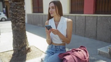 Sarışın, güzel bir kadın, şehir merkezindeki bir bankta oturuyor. Moda kıyafetlerini giyerken telefonunu tutuyor..
