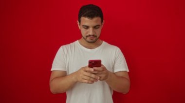 Adam işaret dilini el ve göz ile gösteriyor, genç İspanyol gülümseyen adam izole edilmiş kırmızı arka planda akıllı telefon kullanıyor, mutlu ve kendinden emin bir hareket, parmaklarından neşeyle bakıyor.