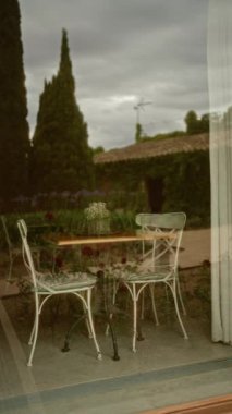 İki beyaz sandalyeli açık hava sahnesi ve bulutlu gökyüzünün altında arka planı olan bir pencereye yansıyan ahşap bir masa.