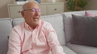 Pembe gömlekli yaşlı bir adam evdeki rahat bir oturma odasında rahat ve rahat bir ortamda rahat bir şekilde oturuyor ve gülümsüyor..