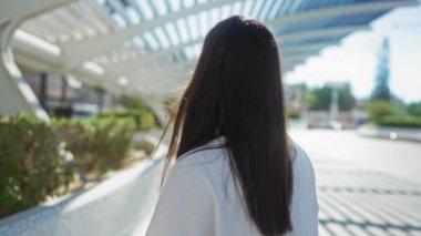 Uzun esmer saçlı bir kadın güneşli bir günde modern mimarisi ve yeşil çalıları olan bir şehir parkında ayakta ve gülümsüyor.