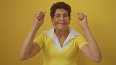 Sarı elbiseli yaşlı İspanyol kadın sarı arka planda izole edilmiş, yumrukları havada ve mutlu bir ifadeyle..
