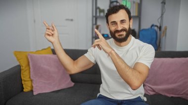 Kapalı alanda gülümseyen sakallı bir adam modern bir oturma odasında el hareketleriyle sağını işaret ediyor..