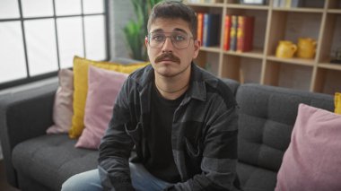 Gözlüklü ve bıyıklı yakışıklı İspanyol bir adam modern bir oturma odasında düşünceli bir şekilde oturuyor..